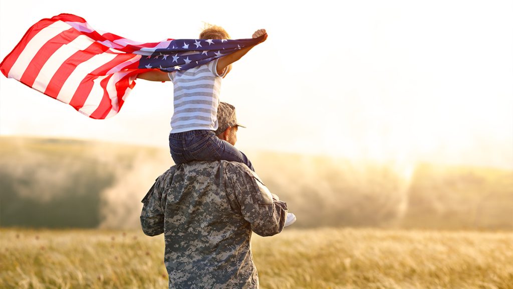 member of the army with a child on their shoulders waving an American flag