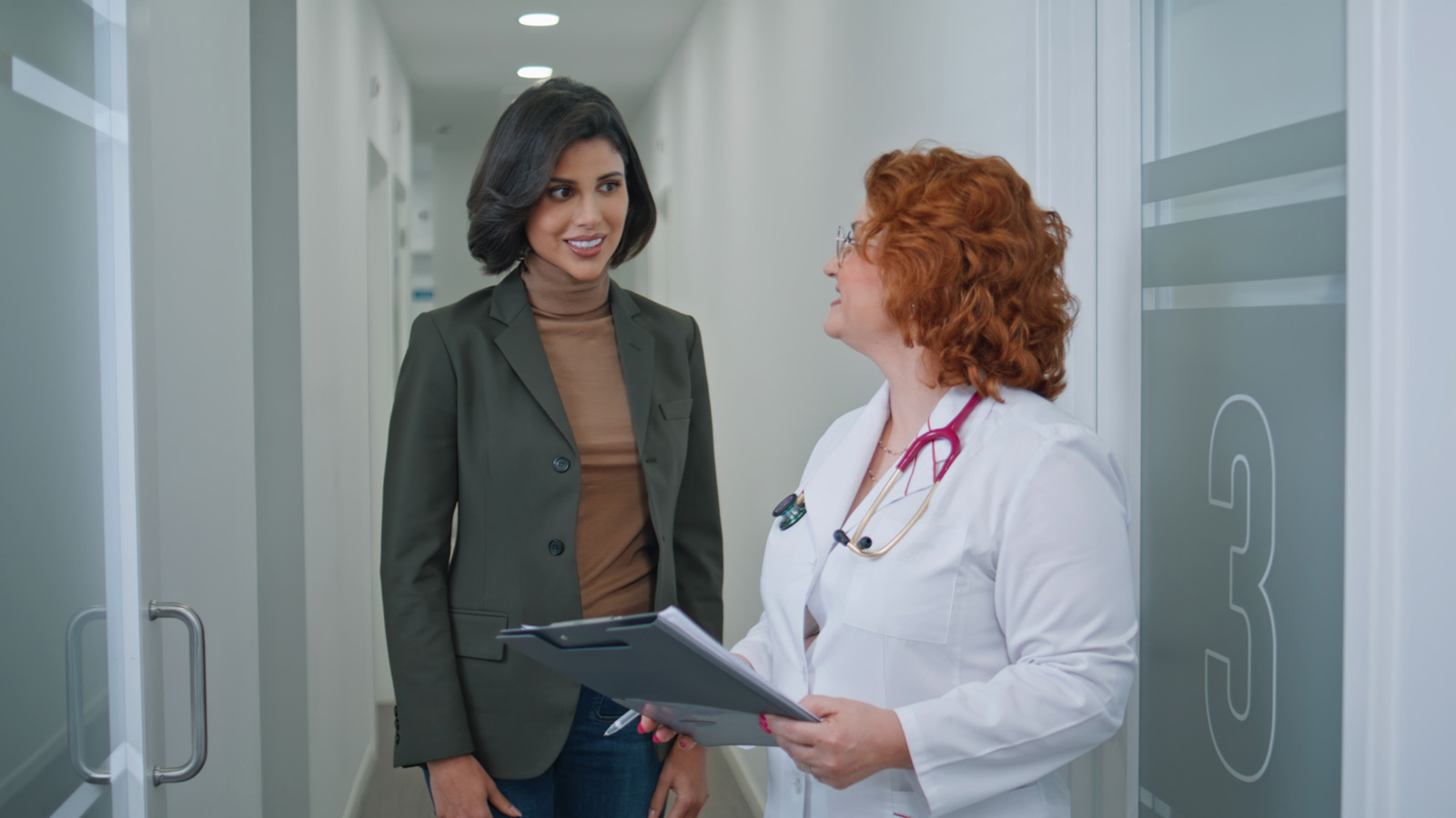 Professional therapist holding clipboard standing with patient in clinic hallway.