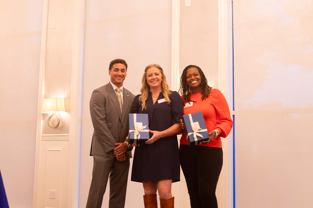 Dean Miller standing and smiling next to two award recipients Abigail Latimer and Shericka Smith, both holding gift boxes