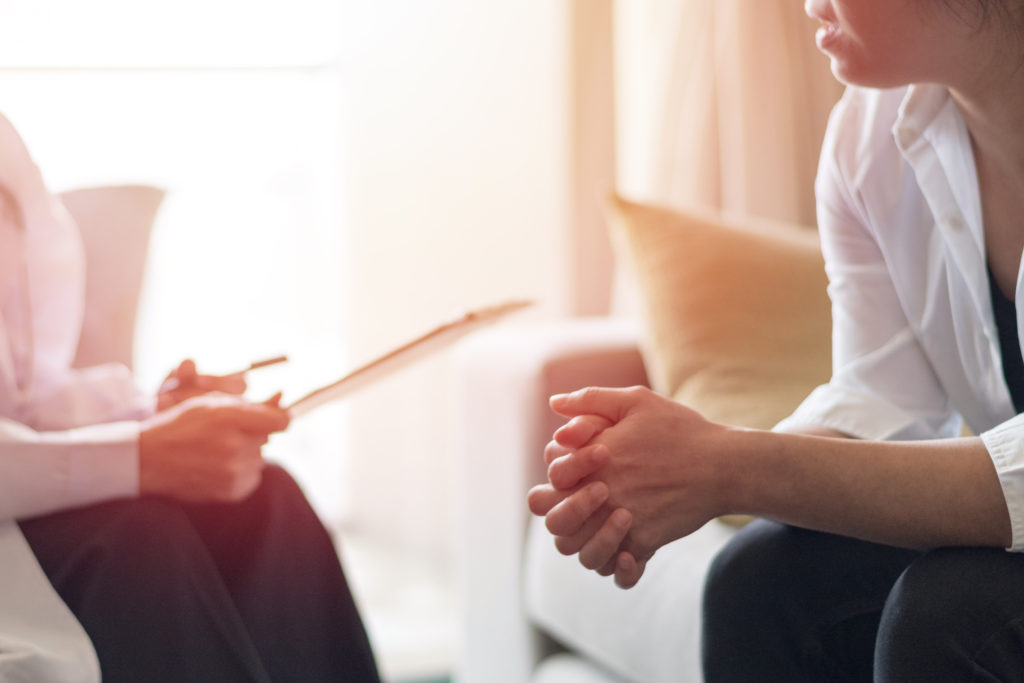Two people sitting in a room, one with a clipboard and one with hands clasped