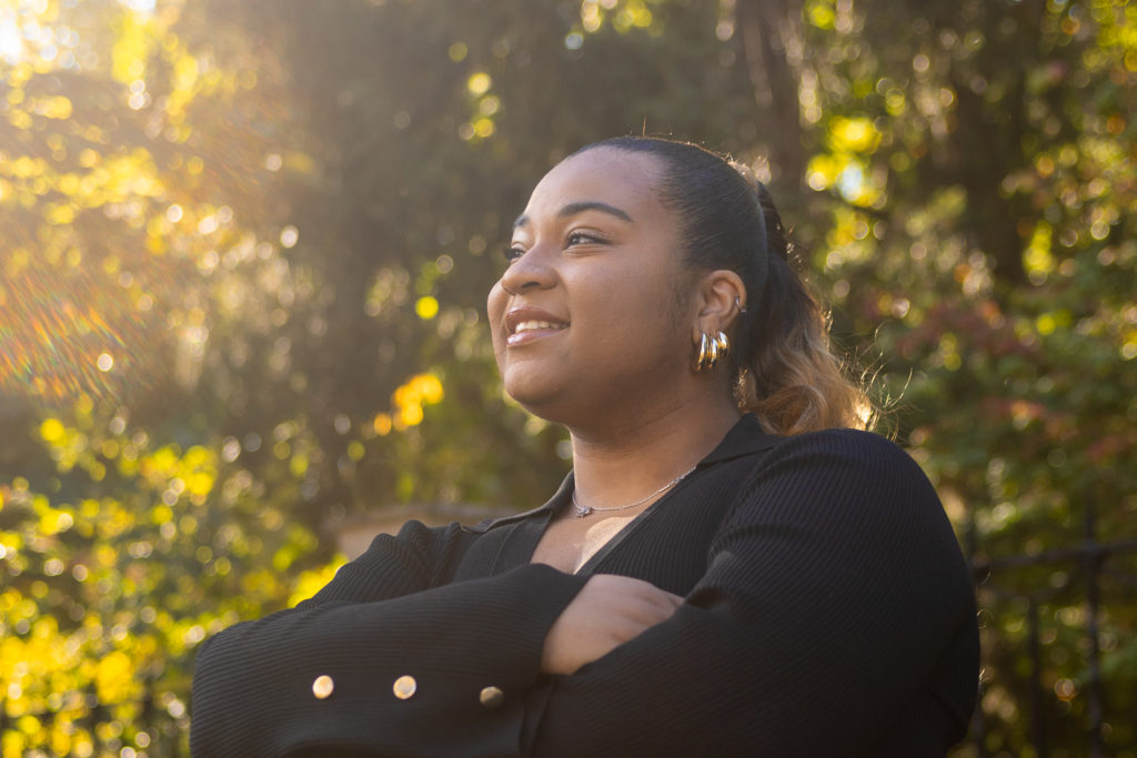 Image of Aniyah Maxey smiling with arms crossed, looking into the distance and smiling with black dress and gold details.