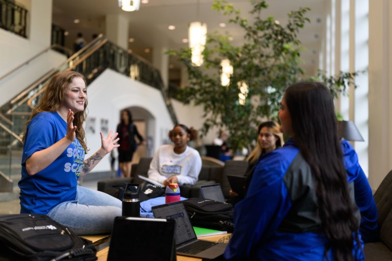 Group of students sitting in a circle and listening to faculty member speak with her arms up