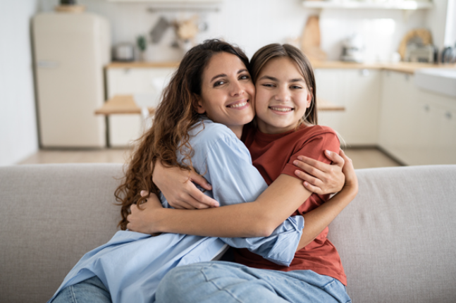 Smiling woman and teenage girl sitting on a couch, hugging each other and looking at the camera.