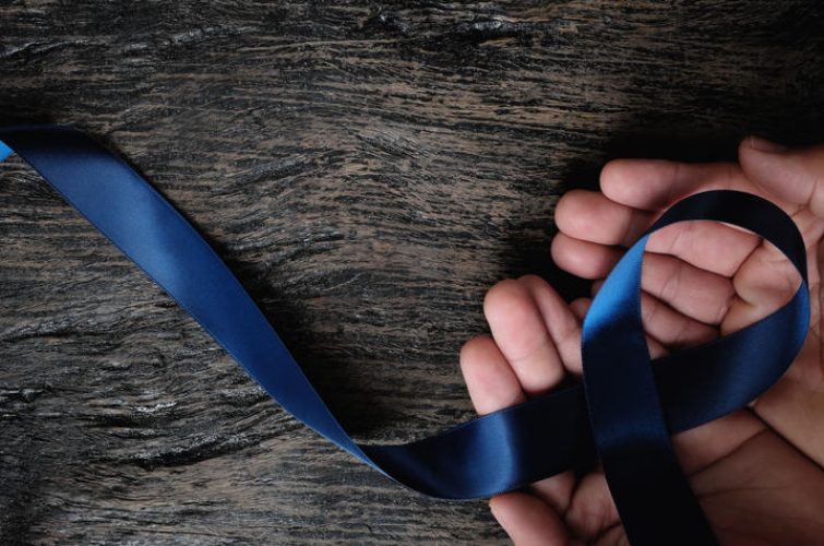 Top view of male hand holding dark blue ribbon on wooden background with copy space. Colon, stomach, colorectal cancer, arthritis, ards acute respiratory distress syndrome awareness and child abuse prevention concept.